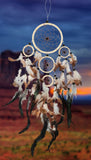 An ivory 5-ring dream catcher comes with a 4-1/2", 2-1/2" and three 1-3/4" rings wrapped in ivory-colored leather adorned with white, brown, and ebony feathers and accented with ebony beads and brown webbing on a blue sunset background.
